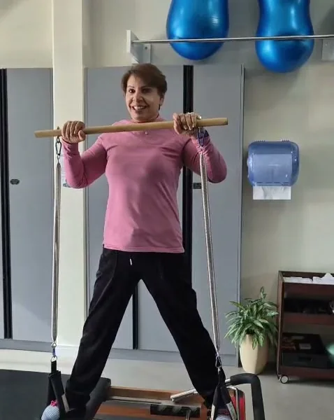 Paciente da melhor idade fazendo exercício em pé no aparelho de Pilates, segurando um bastão